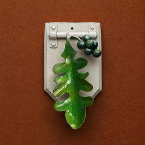 Pode incluir: Um aldravão de metal com uma placa traseira retangular cinza-claro e um mecanismo de dobradiça. Preso à dobradiça está um design de folha de carvalho verde vibrante e um cacho de pequenas bagas verde-escuras, criando uma peça de ferragem.