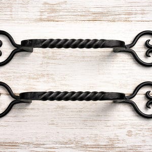 Peut inclure: Deux poignées de porte en fer forgé noir avec un centre torsadé et des extrémités décoratives en volutes. Les poignées sont posées sur un fond en bois clair.