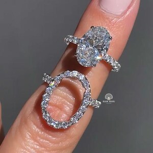 Peut inclure: Deux bagues argentées dotées de montures ovales incrustées de petites pierres claires et scintillantes. La bague supérieure possède une pierre centrale ovale pleine, tandis que la bague inférieure présente un cadre ovale ouvert. Le texte GLAM PEARL est visible.