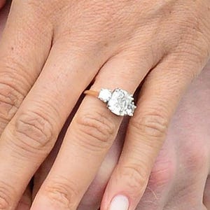 May include: A gold ring with a large, square-cut center diamond and two smaller side stones. The ring is set on a gold band and is on a person's finger. The person is holding another person's hand.