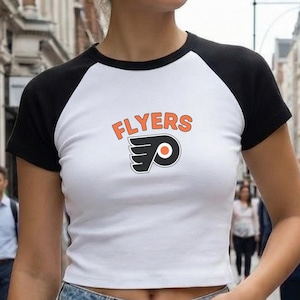 May include: White crop top with black sleeves featuring the word "FLYERS" in orange and a logo. The shirt is paired with blue jeans. The image is taken outdoors on a city street.