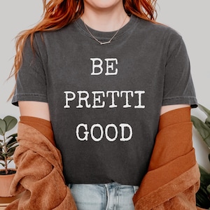 May include: Dark gray t-shirt with the words "BE PRETTI GOOD" in white block letters. The shirt is worn with a rust-colored jacket and blue jeans. A gold side table with a plant is visible in the background.