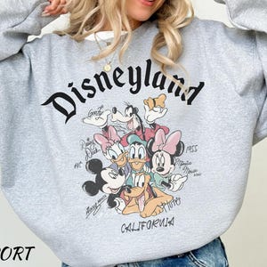 May include: Sport grey sweatshirt featuring the word "Disneyland" in a black script font. The sweatshirt has a graphic of Disney characters including Mickey Mouse, Minnie Mouse, Donald Duck, and Goofy. The word "California" is printed at the bottom.