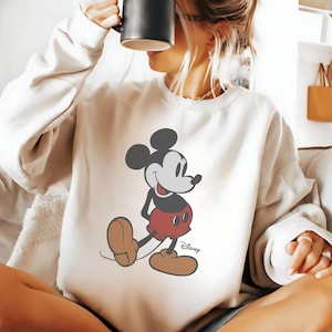 K&ouml;nnte beinhalten: Cremefarbenes Sweatshirt mit einer l&auml;chelnden Mickey Mouse-Grafik. Die Zeichentrickfigur ist in ihrem klassischen Outfit dargestellt: rote Shorts, braune Schuhe und schwarze Handschuhe. Das Disney-Logo ist darunter gedruckt.