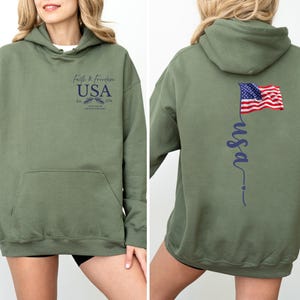 Puede incluir: Sudadera con capucha verde oliva con bolsillo delantero. La parte delantera presenta el texto "Faith & Freedom USA" con un emblema. La parte trasera muestra una bandera estadounidense ondeando y la palabra "usa" en cursiva.