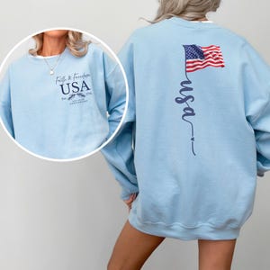 Puede incluir: Sudadera azul claro con la palabra "USA" y "Faith & Freedom" estampadas en la parte delantera. La parte trasera presenta una inscripci&oacute;n vertical "usa" y una bandera americana. La sudadera es de cuello redondo.