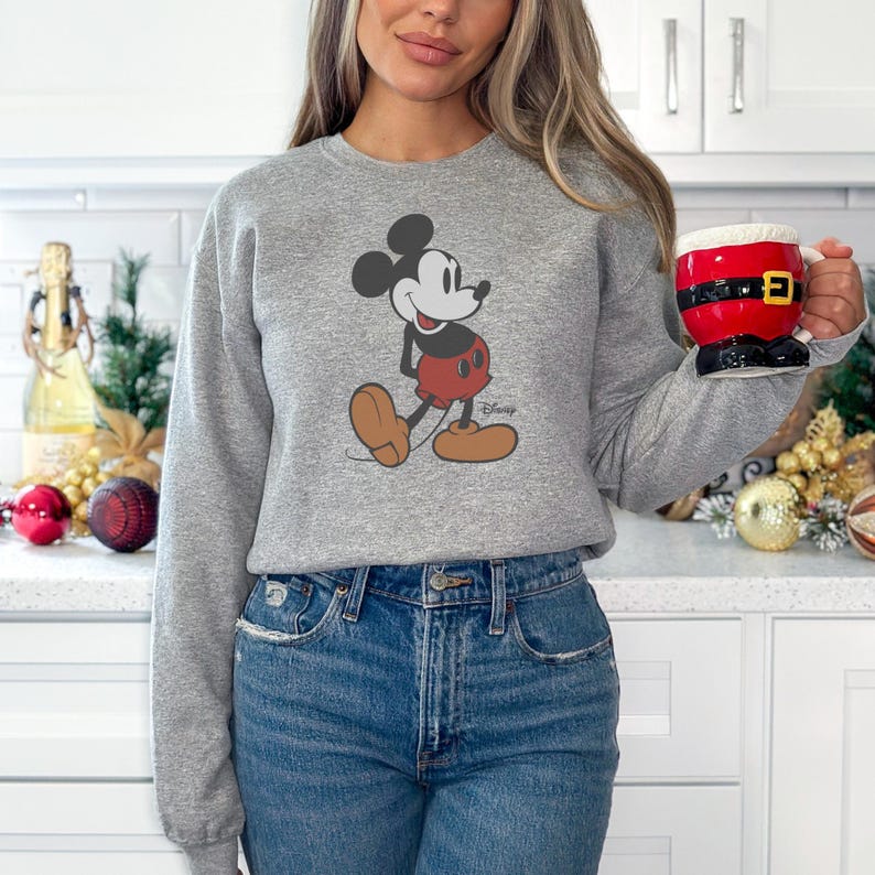 K&ouml;nnte beinhalten: Ein graues Sweatshirt mit einem Mickey Mouse-Design in Schwarz, Rot und Braun. Das Sweatshirt wird mit einer blauen Jeans kombiniert. Eine Weihnachtsmann-Tasse mit schwarzem G&uuml;rtel und goldener Schnalle wird in der Hand gehalten.