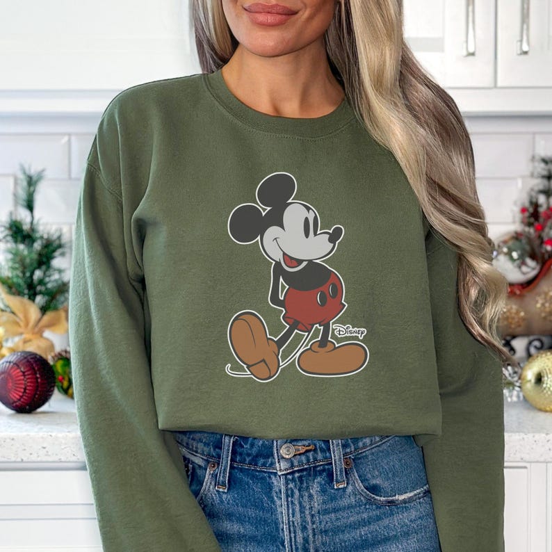 K&ouml;nnte beinhalten: Olivgr&uuml;nes Sweatshirt mit einem Mickey Mouse-Motiv. Die Zeichentrickfigur ist in ihrer klassischen Pose dargestellt und tr&auml;gt rote Shorts und braune Schuhe. Das Disney-Logo ist sichtbar. Das Sweatshirt wird mit blauen Jeans kombiniert.