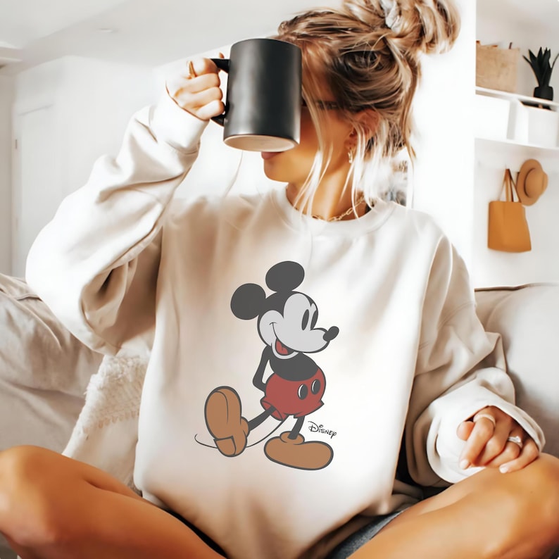 K&ouml;nnte beinhalten: Cremefarbenes Sweatshirt mit einer l&auml;chelnden Mickey Mouse-Grafik. Die Zeichentrickfigur ist in ihrem klassischen Outfit dargestellt: rote Shorts, braune Schuhe und schwarze Handschuhe. Das Disney-Logo ist darunter gedruckt.