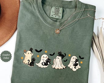 May include: Moss green Comfort Colors t-shirt featuring a Halloween design. The design includes cartoon ghosts, black cats, bats, and stars. The shirt has a crew neck and short sleeves. A silver necklace with a pendant is draped across the shirt.