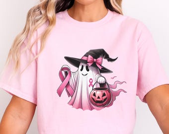 May include: Pink t-shirt featuring a cartoon ghost wearing a witch hat with a pink bow. The ghost holds a pink ribbon and a jack-o'-lantern. The design incorporates pink ribbons, symbolizing breast cancer awareness.