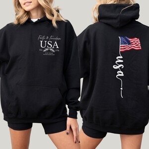 Puede incluir: Sudadera con capucha negra con bolsillo delantero y capucha. La parte delantera presenta el texto "Faith & Freedom USA" con un emblema y "Est. 1776 200 Year Anniversary". La parte trasera muestra una bandera americana ondeando y la palabra "usa" en cursiva.