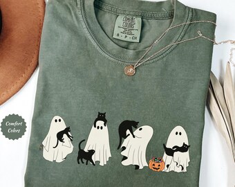May include: Moss green Comfort Colors t-shirt featuring a Halloween design. The design includes four white ghost figures with black cats, and a small orange pumpkin. A silver necklace with a pendant is also visible.