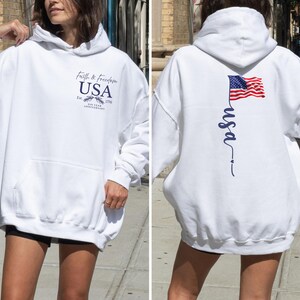 Puede incluir: Sudadera con capucha blanca con bolsillo delantero y el texto "Faith & Freedom USA" con un emblema. La parte trasera presenta una bandera estadounidense ondeando y la palabra "usa" en escritura cursiva. Una prenda casual y patri&oacute;tica.
