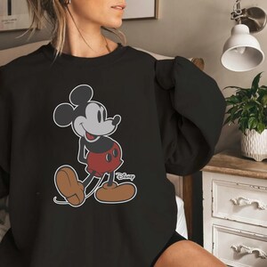 K&ouml;nnte beinhalten: Schwarzes Sweatshirt mit einem Mickey Mouse-Motiv. Die Zeichentrickfigur ist in ihrer klassischen Pose dargestellt und tr&auml;gt rote Shorts und braune Schuhe. Das Disney-Logo ist sichtbar.