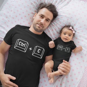 May include: Black t-shirts and a baby onesie with white keyboard key graphics. The adult shirt displays "Ctrl + C", while the onesie shows "Ctrl + V". The items are on a pink and white heart-patterned bedspread.