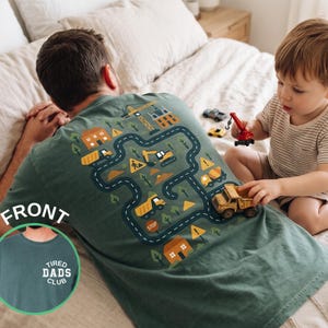 May include: Green t-shirt with a construction site design on the back, featuring roads, buildings, and construction vehicles. The front of the shirt has the text "TIRED DADS CLUB". A child plays with toy construction vehicles.