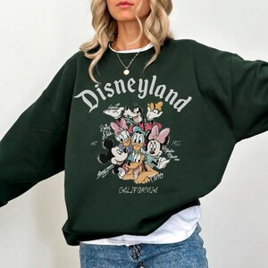 May include: Dark green Disneyland sweatshirt featuring a graphic of Mickey Mouse, Minnie Mouse, Donald Duck, Daisy Duck, Goofy, and Pluto. The word "Disneyland" is arched across the top, with "California" at the bottom.
