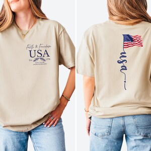 Puede incluir: Camiseta beige con texto "Faith & Freedom USA" y un gr&aacute;fico del 250 aniversario en la parte delantera. La parte trasera presenta una bandera americana ondeando y la palabra "usa" en cursiva.