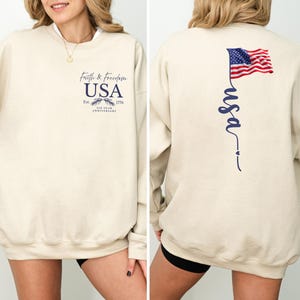 Puede incluir: Sudadera beige con el texto "Faith & Freedom USA" y un gr&aacute;fico de &aacute;guila en la parte delantera. La parte trasera presenta una bandera estadounidense ondeando y la palabra "usa" en cursiva. La sudadera celebra el 200 aniversario.