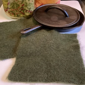 May include: A cast iron skillet with a lid, resting on two olive green knitted pot holders. The skillet has a dark, textured surface and a curved handle. Jars of food are visible in the background.