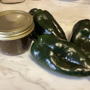 May include: A glass jar with a gold lid filled with a dark brown substance, next to four dark green poblano peppers. The peppers have a glossy sheen and are arranged on a light-colored surface.