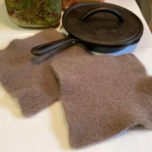 May include: A cast iron skillet with a lid and two brown knitted pot holders. The skillet is black and has a handle. The pot holders are square and made of a soft, textured material. Kitchenware.