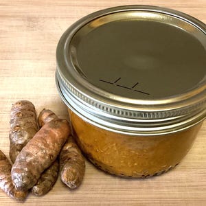 May include: A small glass jar with a metallic lid containing a thick orange paste, positioned next to a cluster of five raw, knobby turmeric roots with a brownish-orange skin. The jar lid features a small, thin, horizontal slit detail.