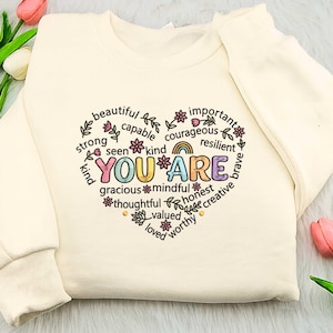 Può includere: Felpa color crema con grafica a forma di cuore. La grafica include le parole "YOU ARE" in lettere colorate, circondate da affermazioni positive come "beautiful", "kind" e "strong". La felpa ha maniche lunghe.