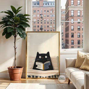 May include: A framed art print featuring a black cat reading a book while reclining on a striped pillow. The artwork has a gold frame and is displayed in a well-lit room with a large potted plant and a white sofa with gold pillows.