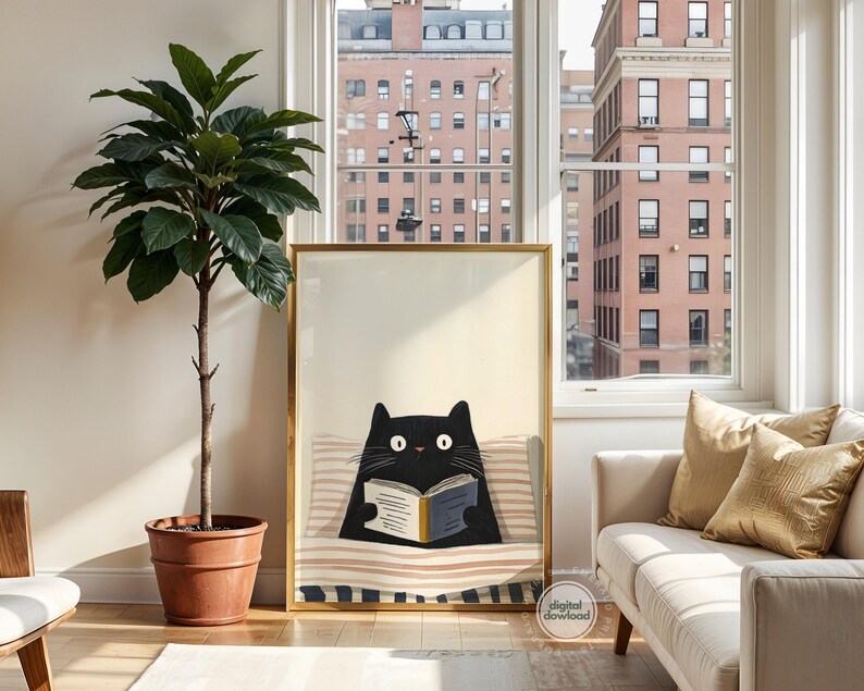 May include: A framed art print featuring a black cat reading a book while reclining on a striped pillow. The artwork has a gold frame and is displayed in a well-lit room with a large potted plant and a white sofa with gold pillows.