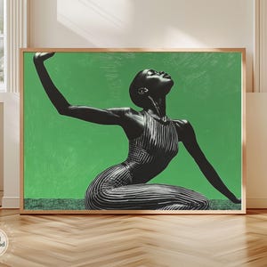 May include: A framed art print featuring a black and white image of a person in a striped dress, kneeling with arms outstretched against a bright green background. The artwork is displayed in a room with natural light.