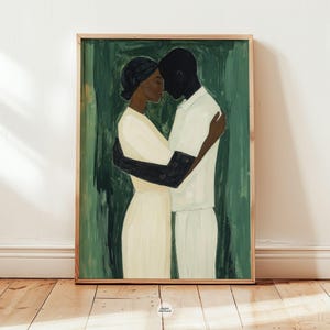 May include: A framed art print depicting two figures embracing. The figures are painted in a minimalist style with a dark-skinned person and a light-skinned person. The background is a textured green, and the frame is a light wood color.