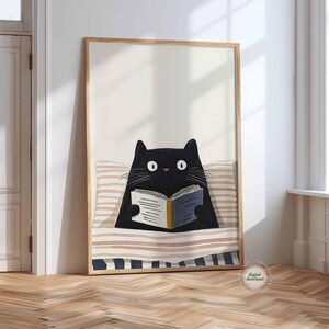 May include: A framed art print featuring a black cat reading a book in bed. The cat has large white eyes and is nestled under a striped blanket. The book has blue and yellow accents. The frame is a light wood color.