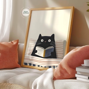 May include: A framed art print featuring a black cat reading a book while lying in bed. The cat has wide eyes and is holding a book with a blue cover. The print is in a wooden frame and is displayed on a sofa with orange pillows.