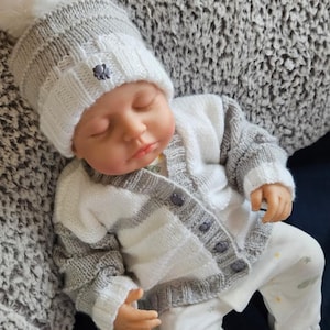 Op de afbeelding: Een babypop met een gebreide grijs-wit gestreepte muts met een witte pompon, een bijpassend vest en een witte voetpyjama. De pop ligt op een grijs oppervlak met textuur.