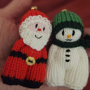May include: Two knitted Christmas ornaments: Santa Claus and a snowman. The Santa ornament is red with black stripes, a white beard, and a gold buckle. The snowman is white with a green hat and scarf, and black button details.