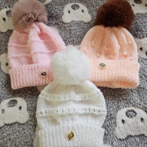 May include: Three knitted baby hats in pink, peach, and white, each topped with a fluffy pom-pom. The pink hat has a mauve pom-pom, the peach hat has a brown pom-pom, and the white hat has a white pom-pom. Each hat has a decorative button.
