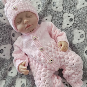 Può includere: Una tutina per neonati in maglia rosa chiaro con berretto abbinato dotato di pon-pon. La tutina presenta un motivo a bolle in rilievo, maniche lunghe e una chiusura frontale con piccoli bottoni decorativi. Il berretto ha un bottone abbinato.