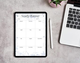 以下が含まれることがあります： 花柄のアクセントが付いた「年間プランナー」レイアウトを表示するデジタルタブレット。プランナーには月ごとのセクションと年のスペースが含まれています。白いスタイラスがタブレットの横にあり、背景にはラップトップと小さな鉢植えの植物があります。