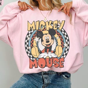Puede incluir: Sudadera rosa claro con un gr&aacute;fico vintage de Mickey Mouse. El dise&ntilde;o incluye las palabras "Mickey Mouse" en una fuente retro, sobre un fondo circular con estrellas y un patr&oacute;n de tablero de ajedrez. La sudadera es de estilo casual.