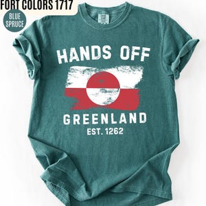 May include: Teal-colored t-shirt with the words "HANDS OFF GREENLAND" in white block letters. The shirt features a distressed Greenland flag design in red, white, and a red circle. The text "EST. 1262" is below the flag.