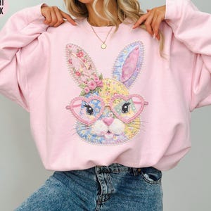 May include: Light pink sweatshirt featuring a colorful bunny face with heart-shaped glasses. The bunny design includes floral and patchwork details. The sweatshirt is a soft, casual style.