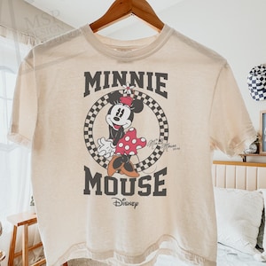 May include: Ivory-colored boxy tee featuring a graphic of Minnie Mouse. The design includes Minnie in a polka-dot dress and a checkered circle with the words "MINNIE MOUSE" and "Disney". The shirt is hanging on a wooden hanger.