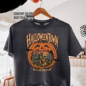 May include: A charcoal gray boxy t-shirt featuring a graphic print of a large carved pumpkin with a town scene inside. The text reads HALLOWEENTOWN and EST. 1998. The shirt is displayed on a wooden hanger against a neutral background.