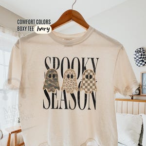 May include: An ivory boxy t-shirt featuring the black text SPOOKY SEASON and three ghost illustrations. The ghosts have different patterns: one with camouflage, one with leopard print, and one with a tan and brown checkerboard pattern.
