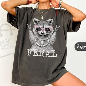 May include: Dark gray t-shirt featuring a raccoon wearing pink sunglasses and a pearl necklace, with the word "FERAL" in white block letters. The raccoon is making a rock and roll hand gesture. The shirt has a relaxed fit.