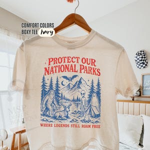 May include: An ivory boxy t-shirt featuring a graphic print in red and blue. The text reads "PROTECT OUR NATIONAL PARKS" and "WHERE LEGENDS STILL ROAM FREE." The illustration depicts mountains, trees, a bat, a bigfoot, a deer, an alien, and a ghost.