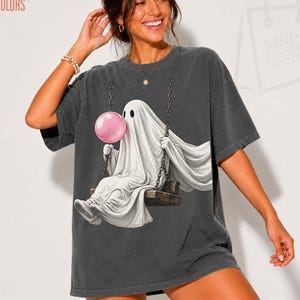 May include: A charcoal gray short-sleeve t-shirt featuring a graphic print of a white ghost sitting on a wooden swing. The ghost is holding a large pink bubblegum sphere. The shirt has a crew neckline and a relaxed, loose-fitting silhouette.