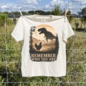 May include: A cream-colored short-sleeve t-shirt featuring a graphic print of a chicken standing in the foreground with a large dinosaur silhouette in the background. The text REMEMBER WHO YOU ARE is printed in a dark brown font below the graphic.